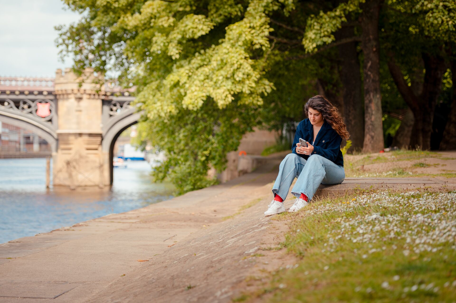 A student uses a phone as she sits on the bank of a river